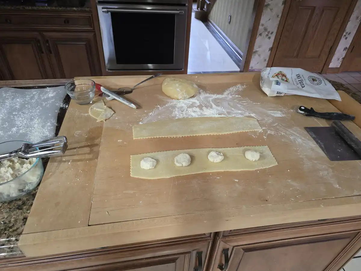 A kitchen counter shows pasta-making in progress with dough rolled out, four dollops of filling placed in a line on one…