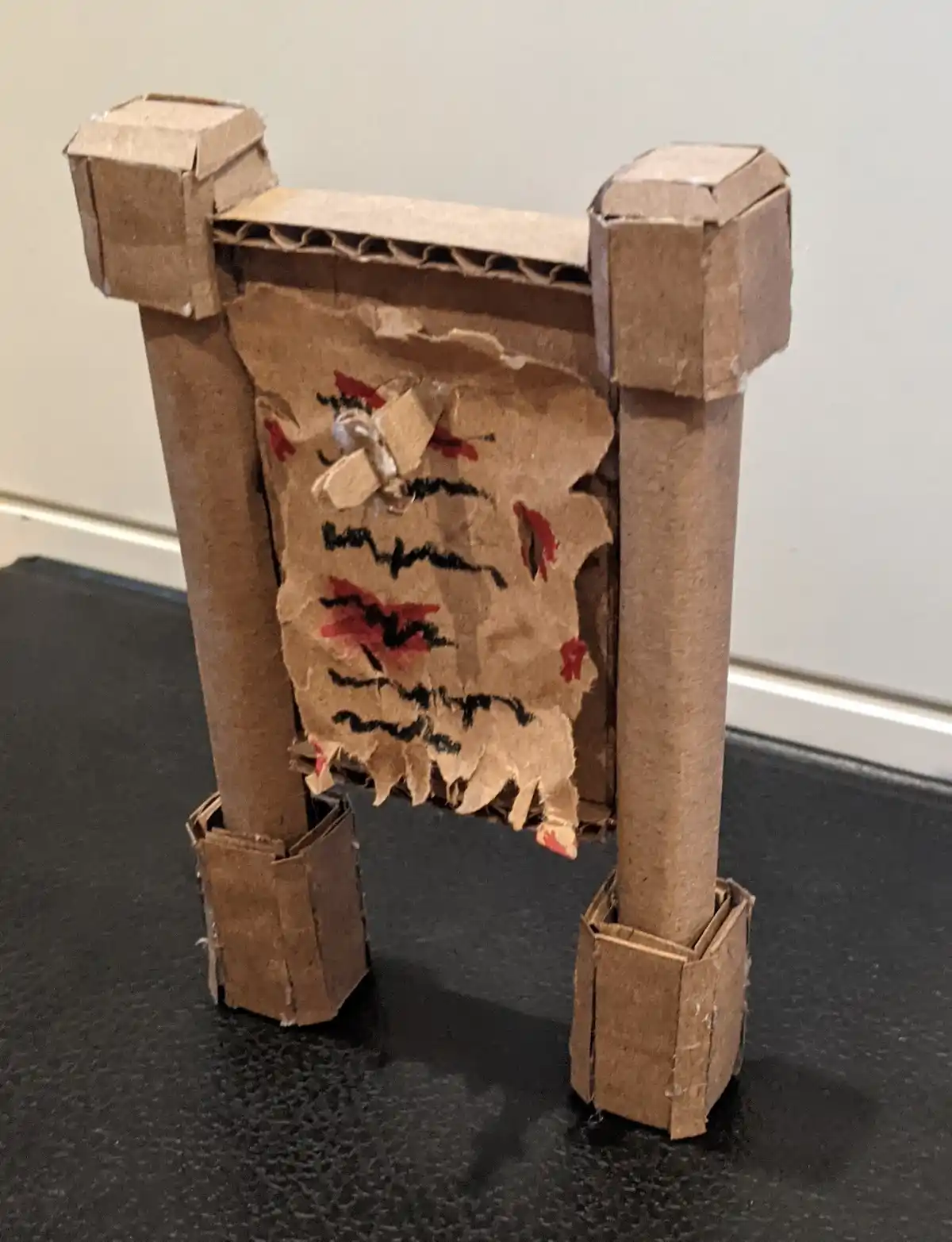 A handmade cardboard gate with two vertical pillars and a horizontal top, standing on a dark surface. The gate has a torn…