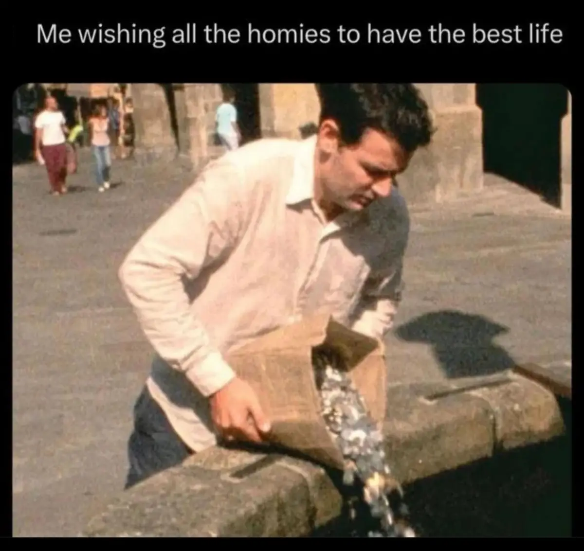 A man in a light-colored shirt is pouring coins from a brown paper bag into a stone water fountain in a public square. Other…