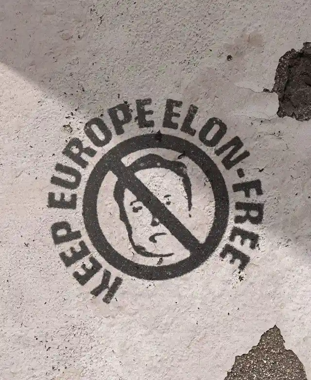 A black stencil graffiti on a weathered, cracked concrete wall shows a circular 'no' symbol over a crude face sketch, with…