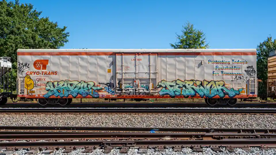 A white and orange Cryo-Trans refrigerated railcar sits on tracks under a clear blue sky, covered in colorful graffiti. The…