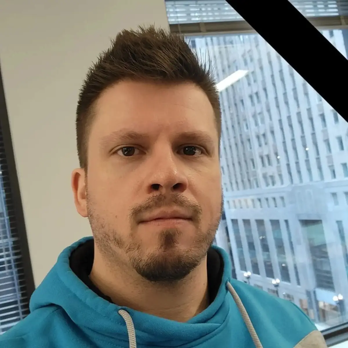 A man with short brown hair and a goatee, wearing a blue hoodie, stares directly at the camera with a neutral expression…