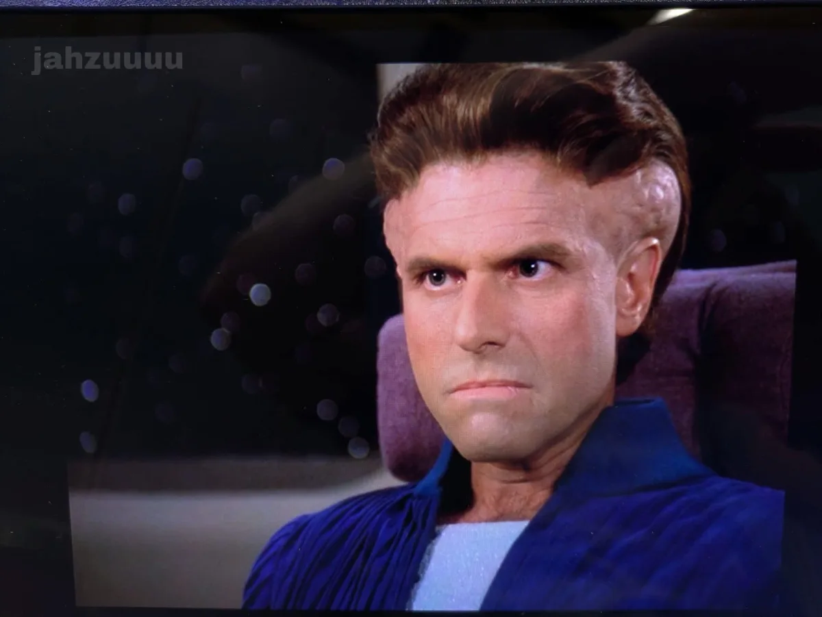 A close-up of a man with a stern, unimpressed expression, wearing a blue robe over a white shirt, seated in a purple chair…