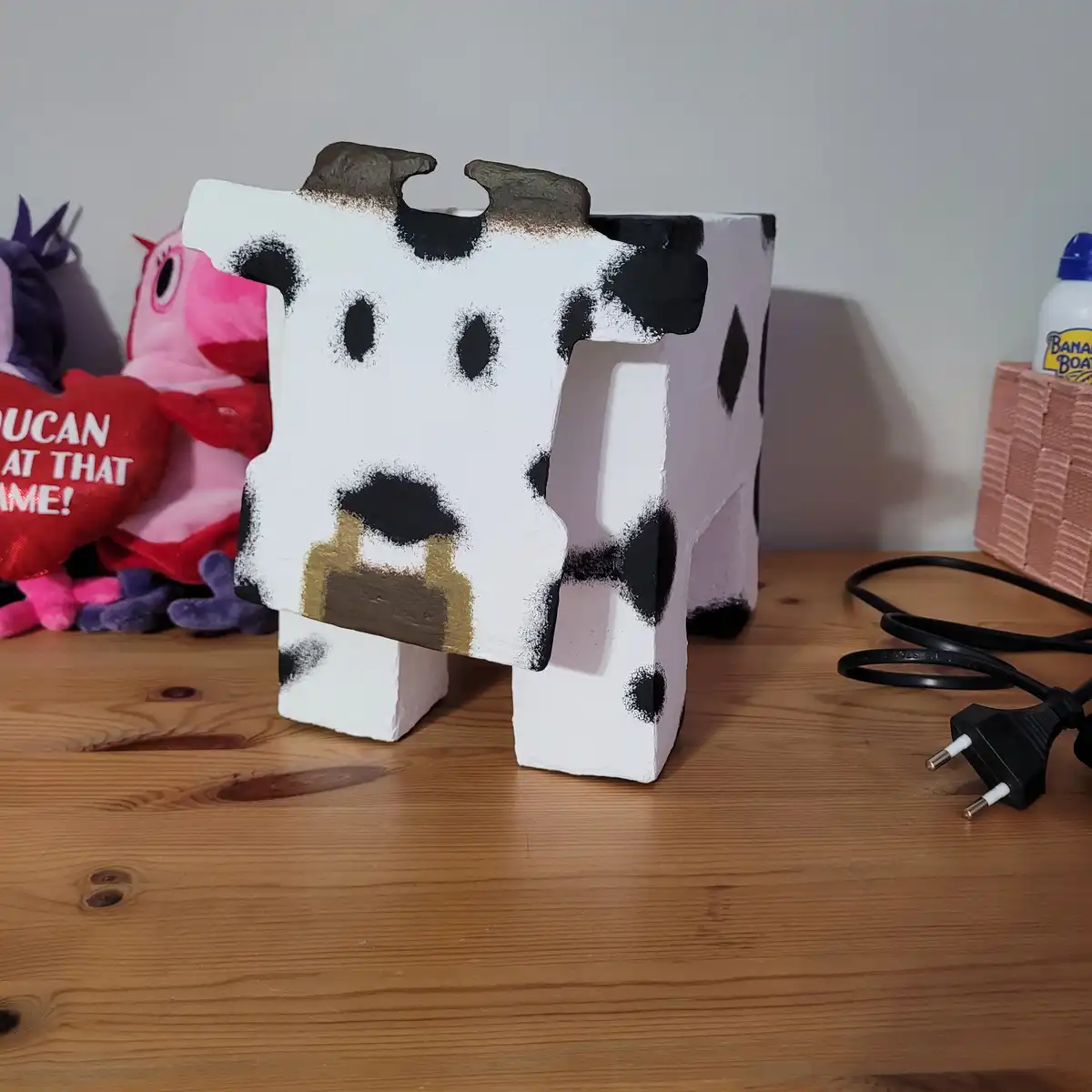 A blocky, cube-shaped cow figurine with black spots and a brown snout sits on a wooden table, next to a coiled black power…
