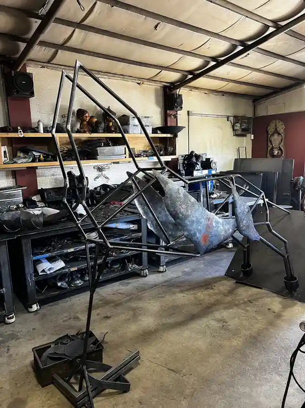 A metal sculpture of a whale skeleton is partially assembled in a cluttered workshop, surrounded by tools and materials. The…