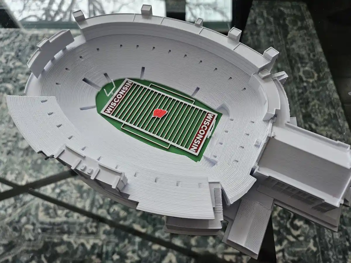 A 3D-printed model of a football stadium with a green field and white bleachers, featuring the word 'WISCONSIN' on both ends…