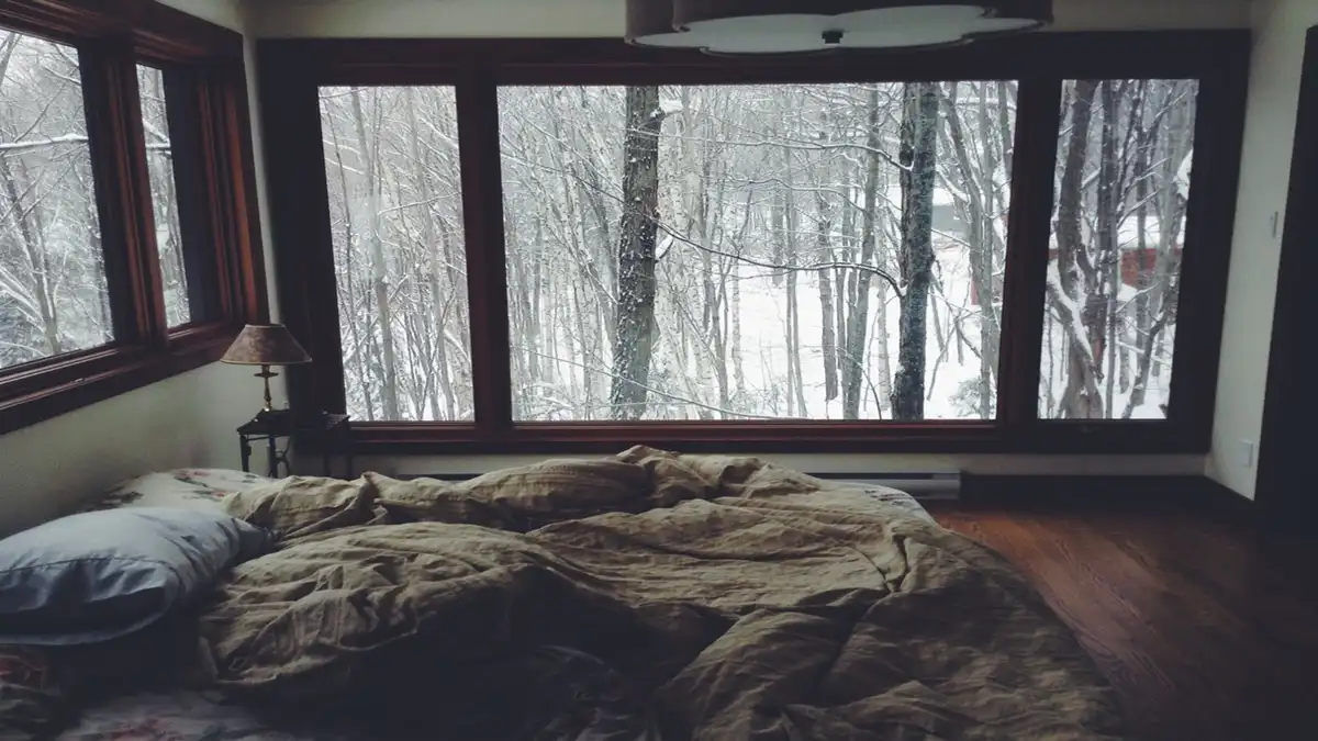 A cozy, messy bedroom with an unmade bed and rumpled blankets, featuring large windows overlooking a snowy forest. The…