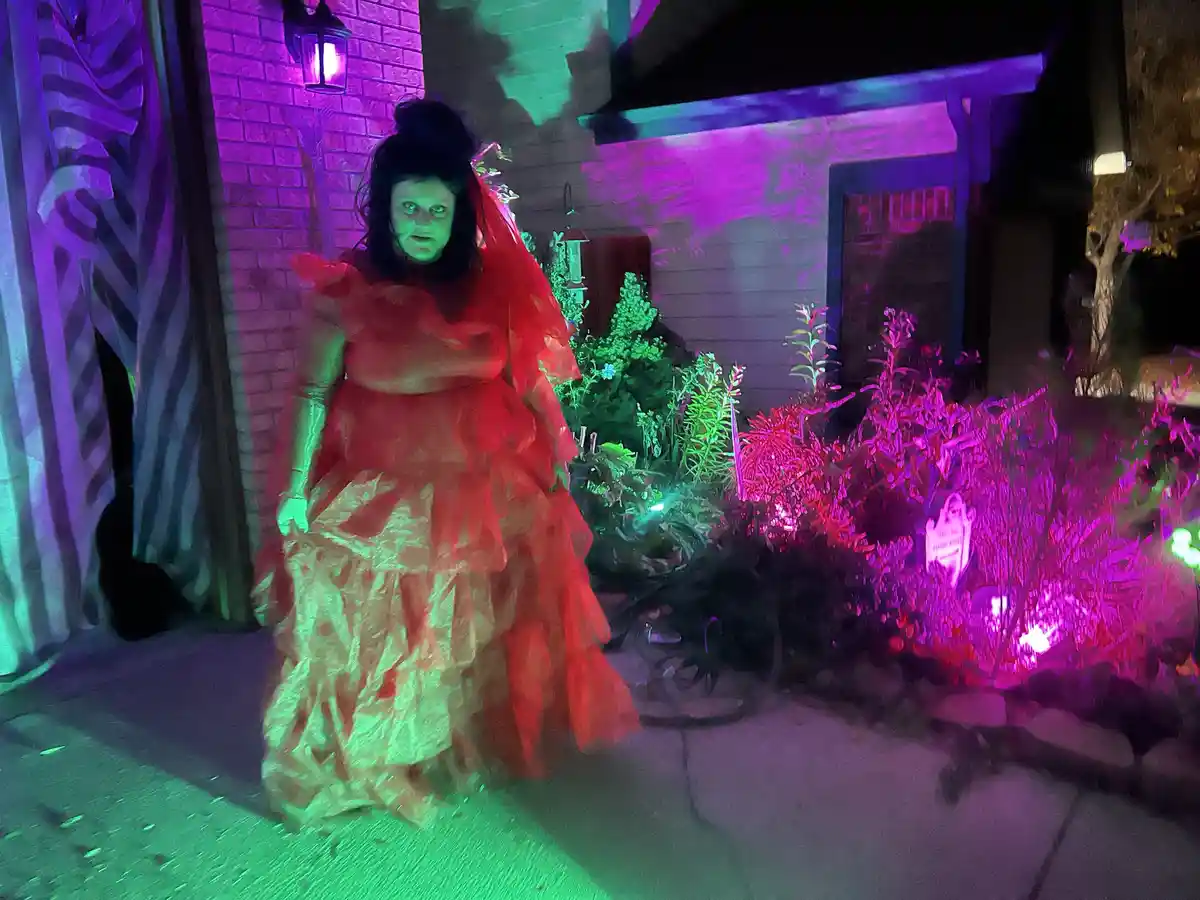 A person in a red, ruffled, layered costume with a veil and pale green face paint stands on a sidewalk in front of a house…