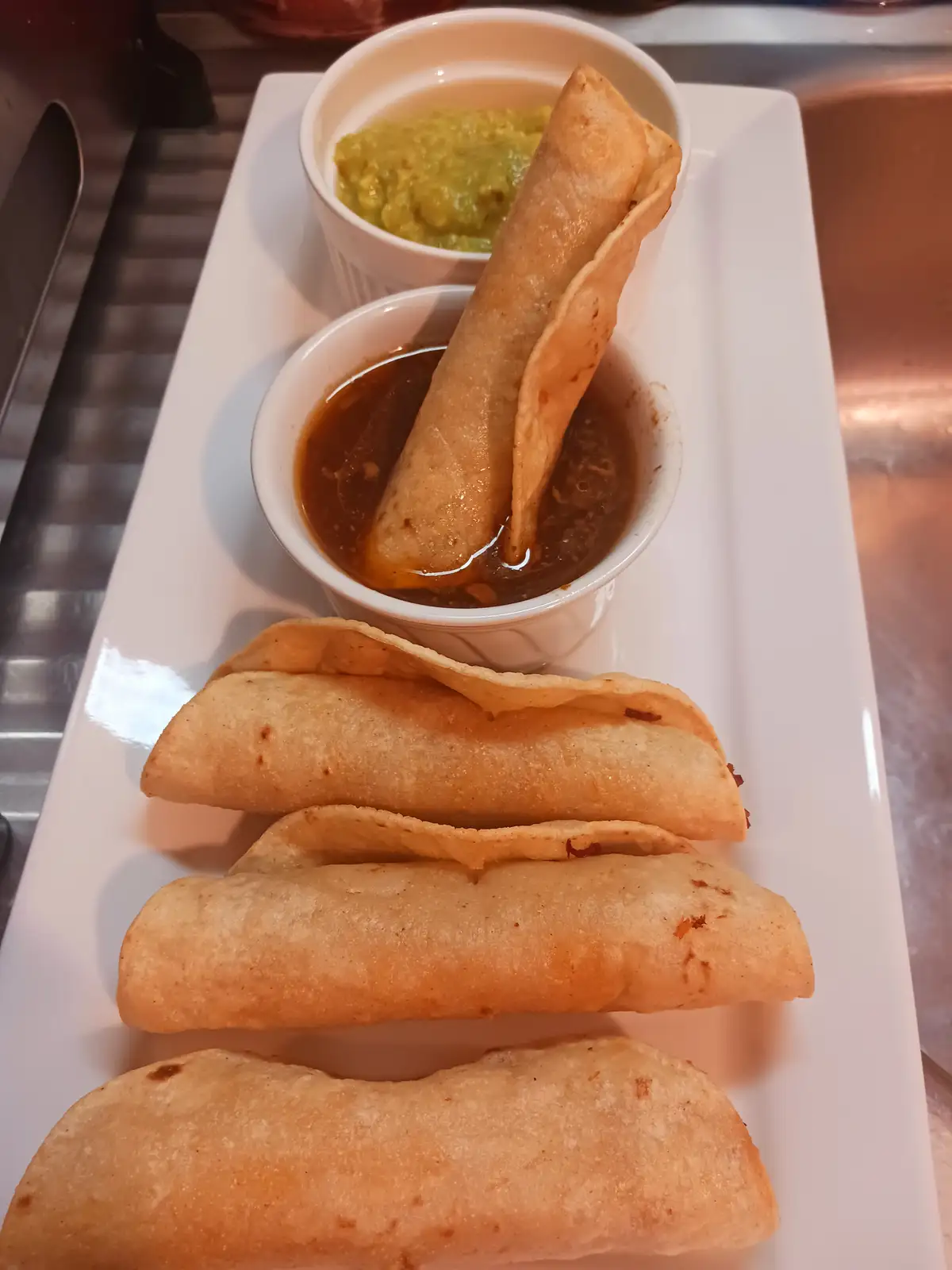 A white rectangular plate holds four golden-brown fried taquitos arranged in a row. One taquito is dipped into a small white…