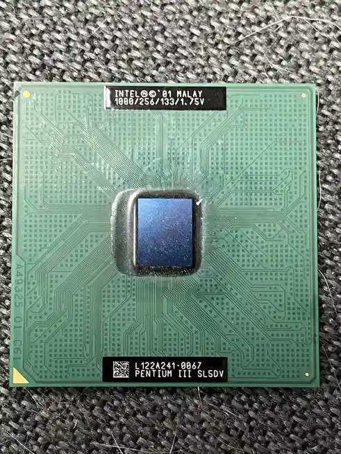 A close-up photo of an Intel Pentium III CPU die on a green circuit board, resting on a textured gray fabric. The chip has…