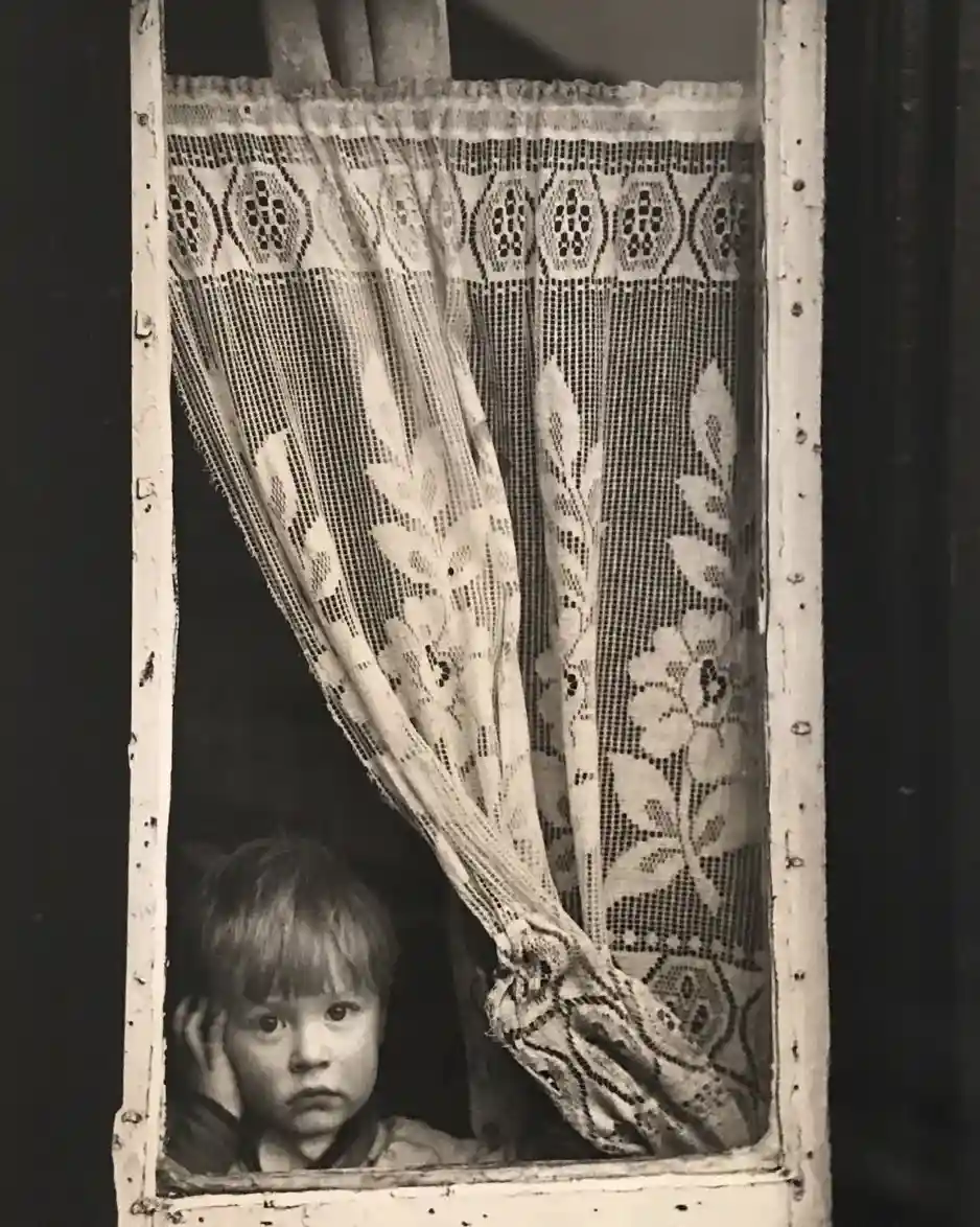 A black-and-white photograph shows a young child peeking out from behind a lace curtain in a weathered window frame, looking…