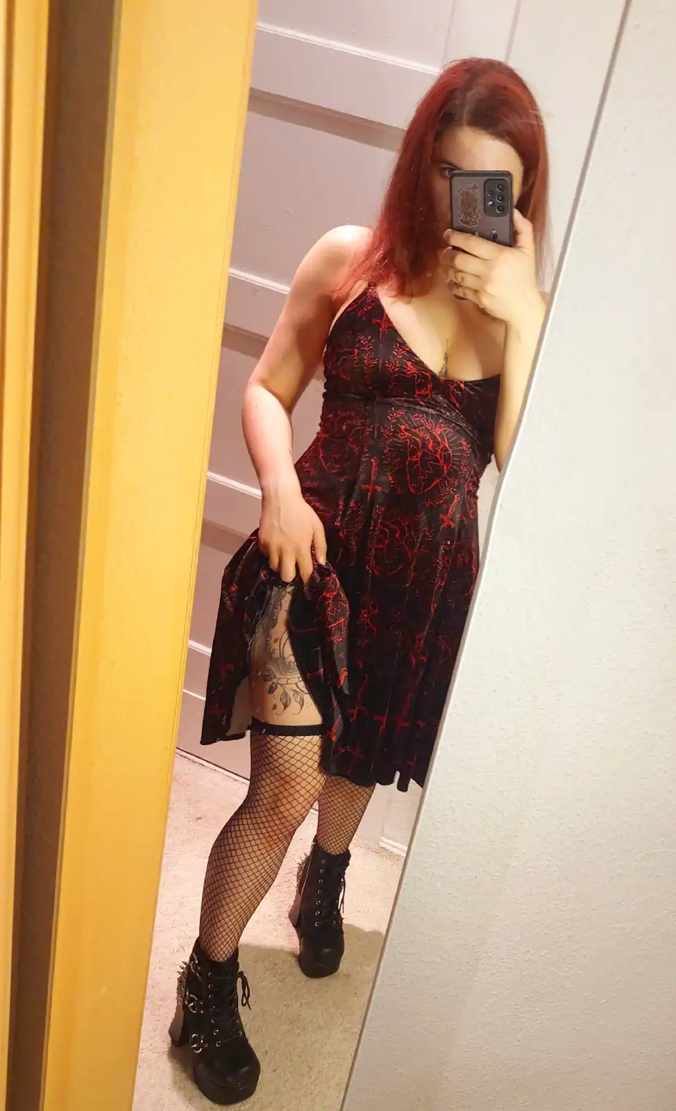 A person with red hair takes a mirror selfie while wearing a black dress with red patterns, fishnet stockings, and black…