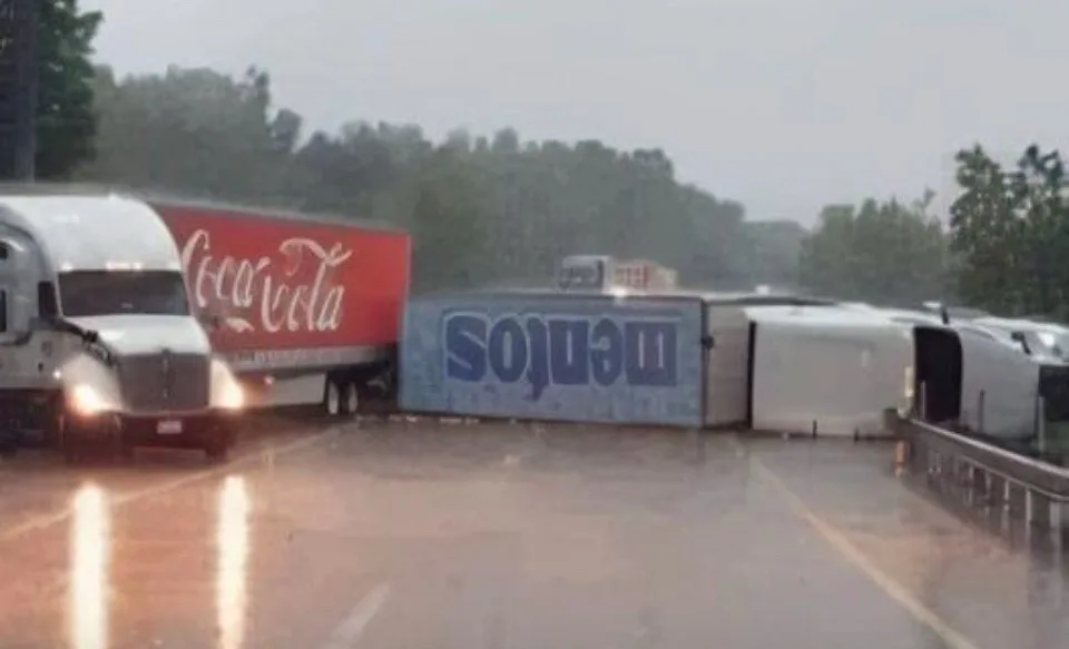A rainy highway scene shows a Coca-Cola branded semi-truck driving past a severely overturned and damaged Melos-branded…