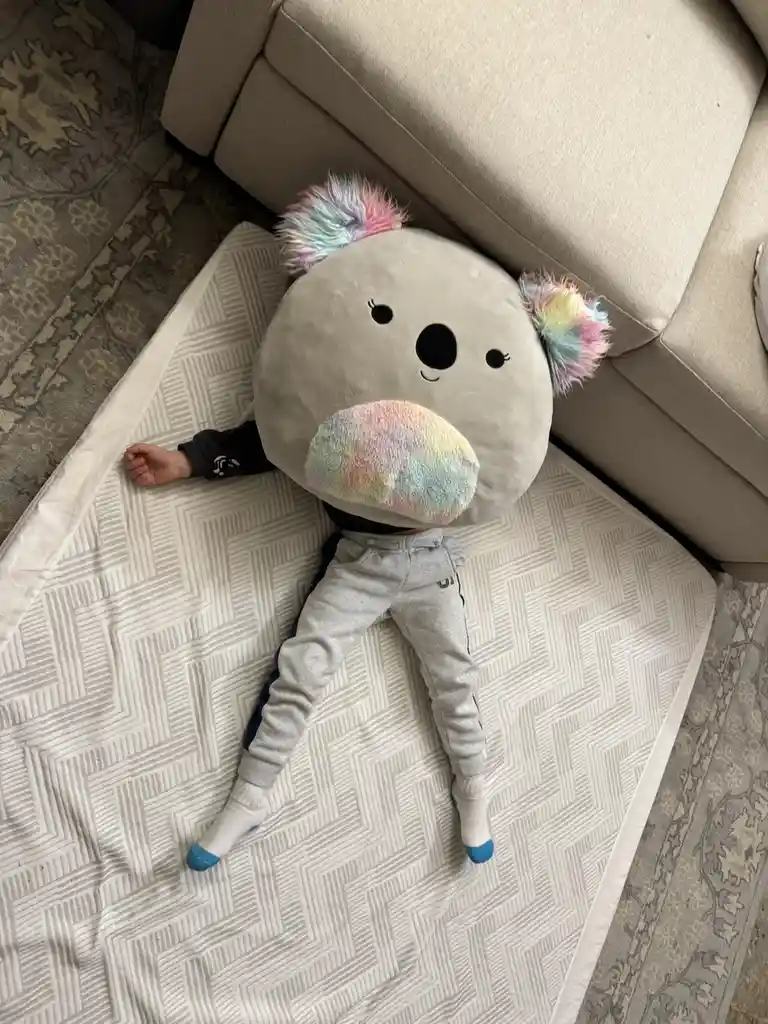 A child is lying on their back on a patterned mat, with their head and upper torso obscured by a large, round, grey koala…