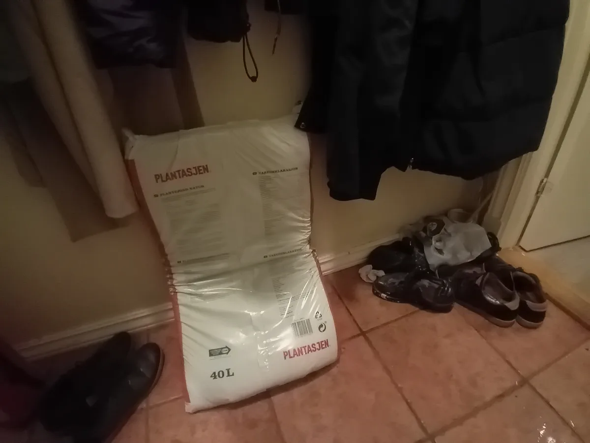 A large white bag labeled 'PLANTASJEN' and '40 L' sits on a tiled floor in a hallway, surrounded by shoes and hanging…