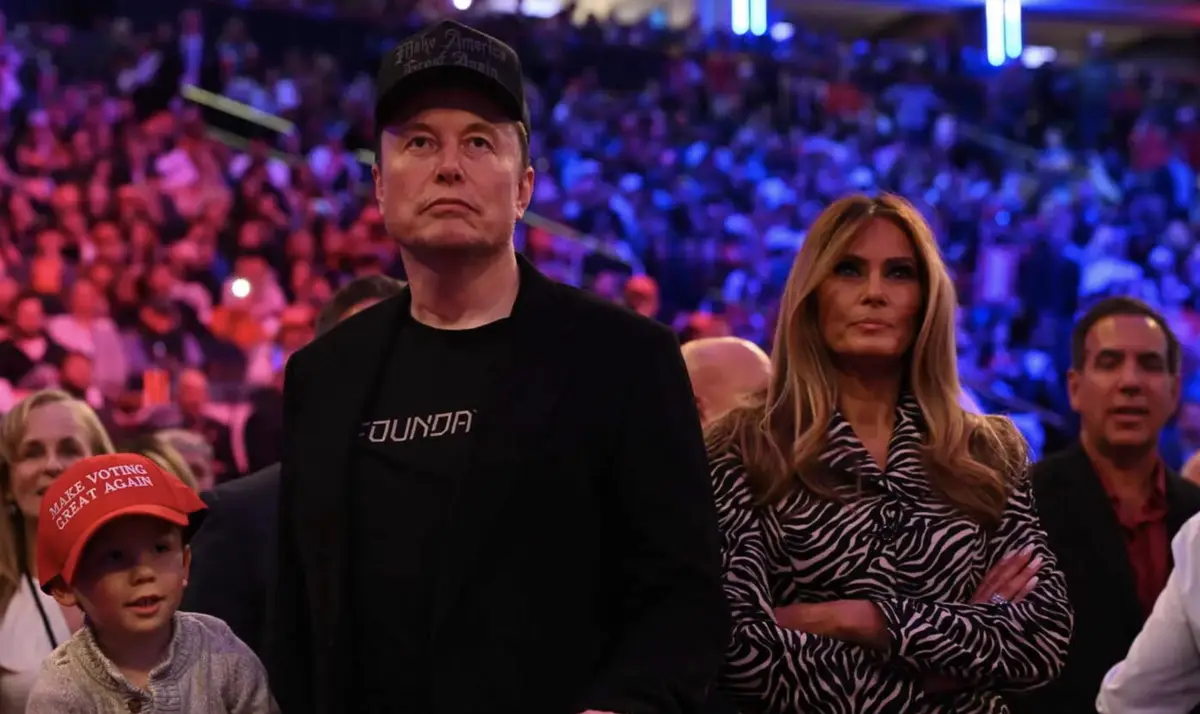 elon musk wears a 'make america great again' cap while sitting in a crowd, next to melania trump who has her arms crossed. a…