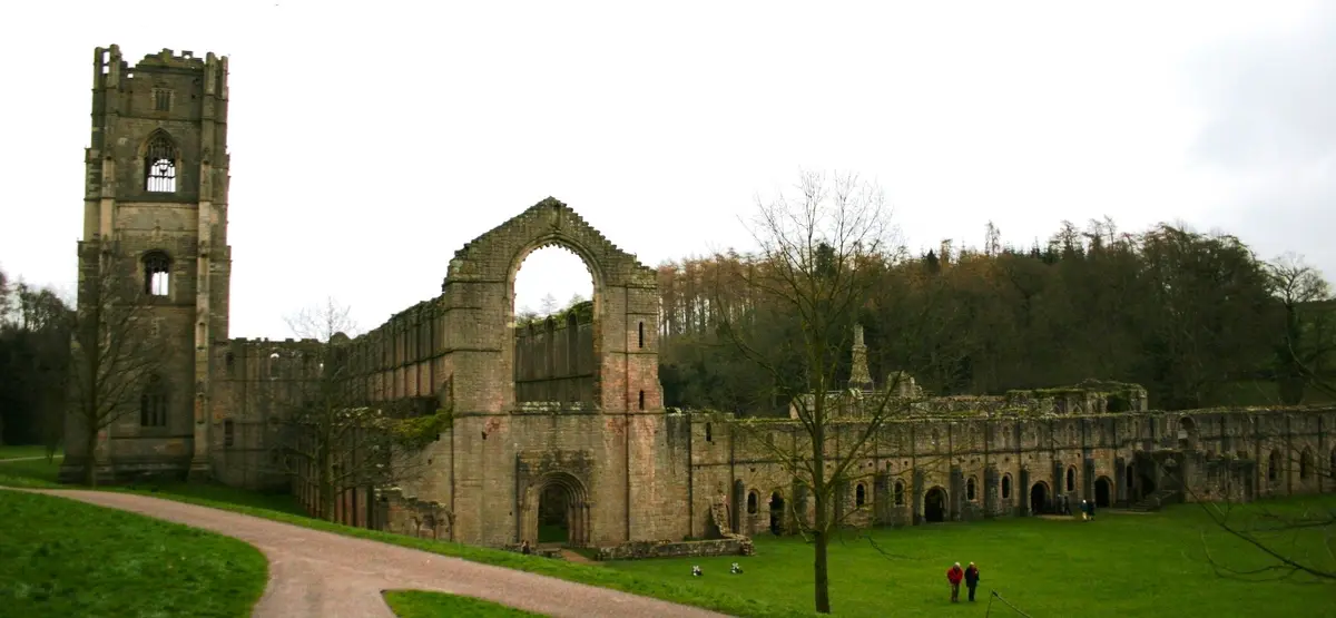 The image shows the ruins of a large, ancient stone abbey with a prominent tower and arched windows, set against a cloudy…