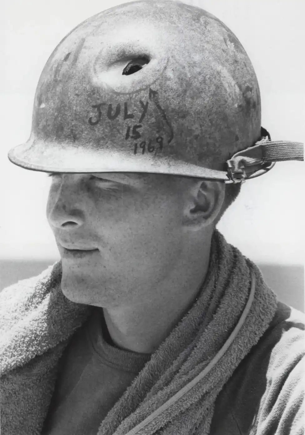 A black and white photo shows a young man in profile wearing a military helmet with 'july 15 1969' written on it. He has a…