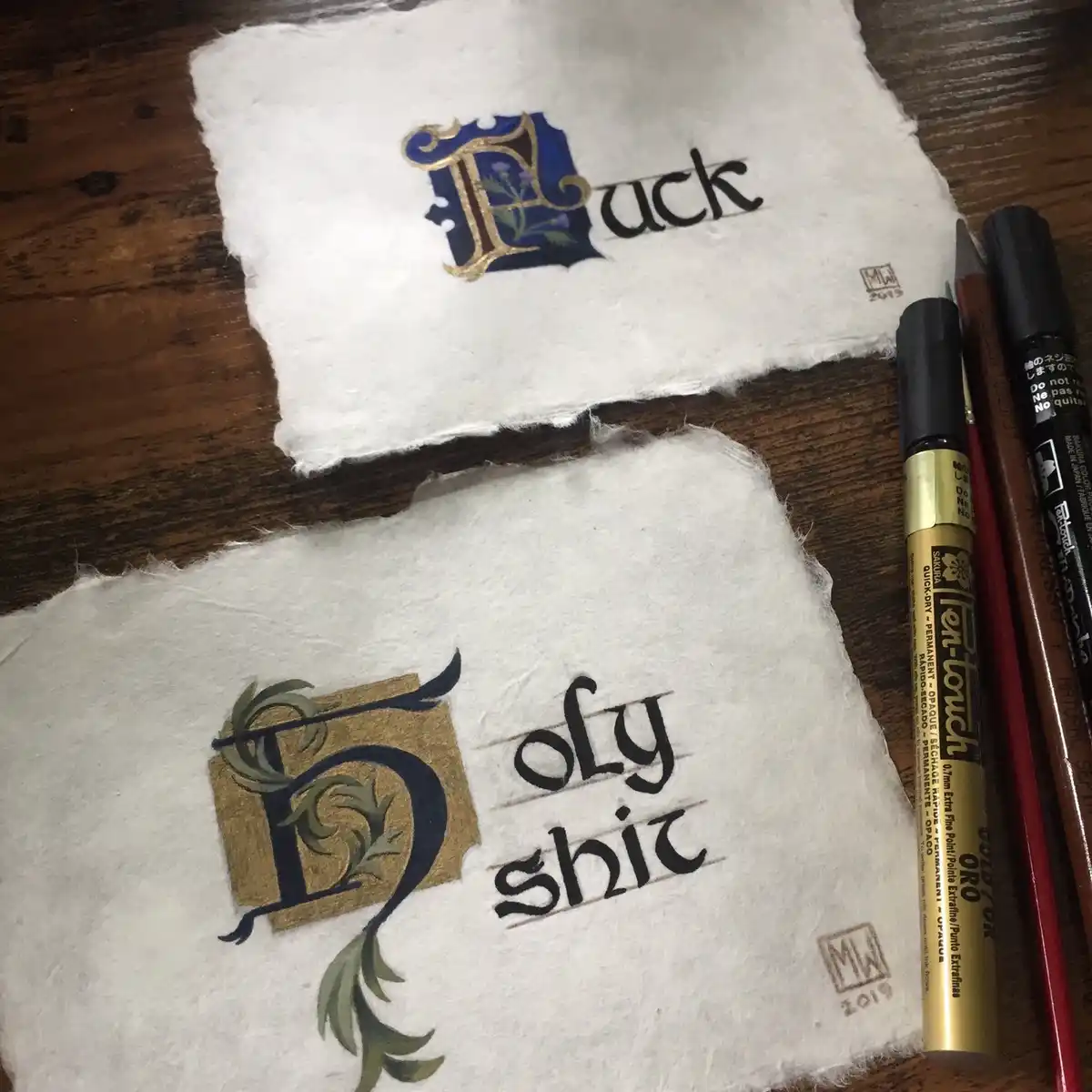 Two pieces of handmade, textured white paper on a wooden surface, each featuring ornate, calligraphic lettering. The top…