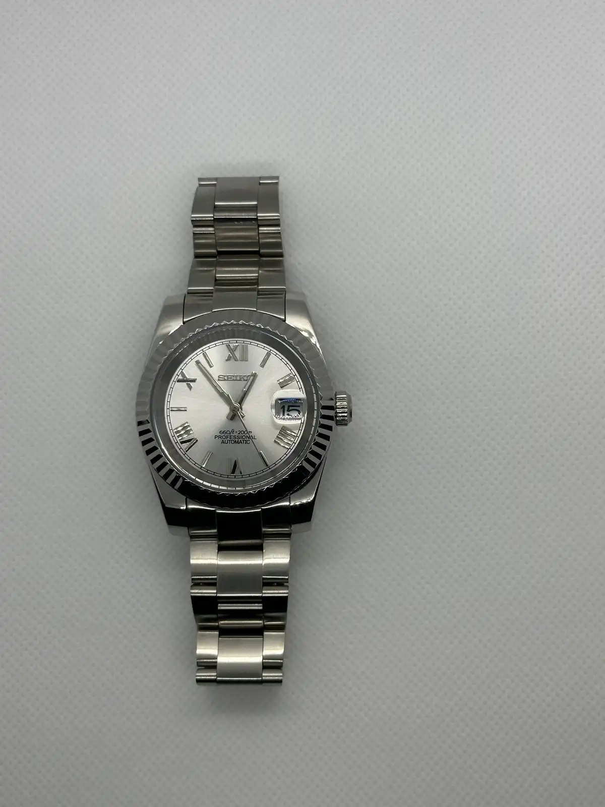 A silver Seiko watch with a stainless steel bracelet and Roman numeral dial is photographed on a textured gray surface. The…