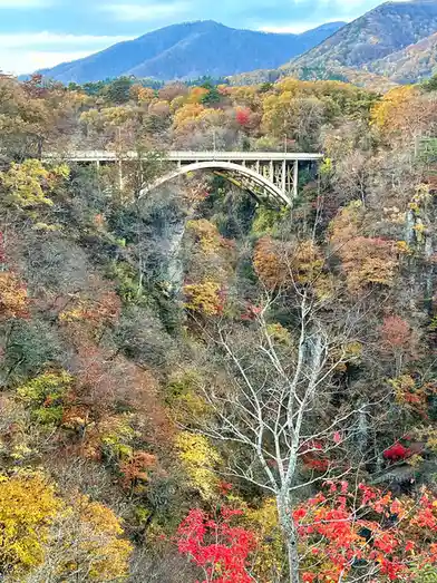 A scenic autumn photo shows a large arched bridge spanning a deep valley surrounded by mountains covered in vibrant red…