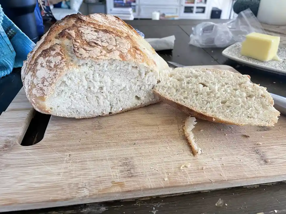 A freshly baked, rustic loaf of bread with a golden-brown crust sits on a well-used wooden cutting board, one slice cut and…