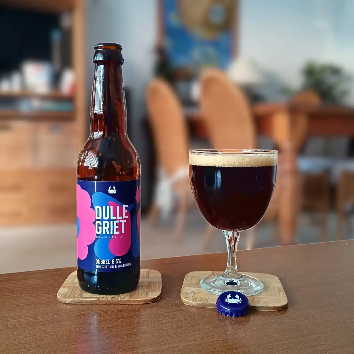 A brown beer bottle labeled 'DULLE GRIET' sits next to a glass of dark beer on a wooden table, both resting on coasters. The…