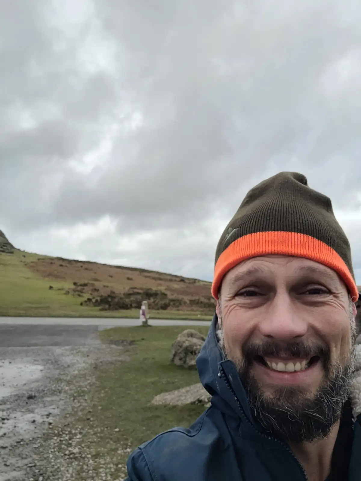 A man with a beard and a brown beanie with an orange stripe smiles broadly at the camera while standing outdoors in a…