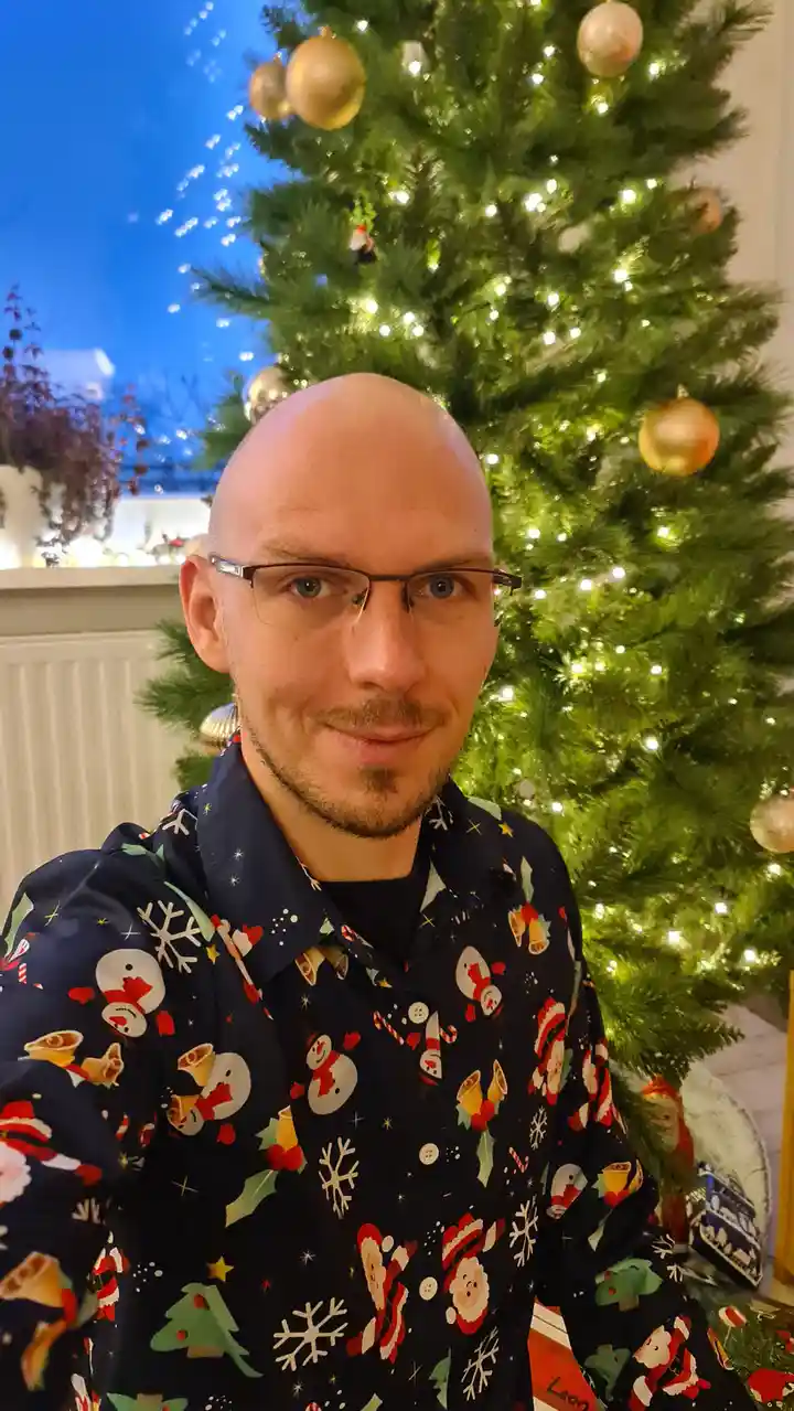 A bald man with glasses and a goatee wears a dark blue shirt covered in festive Christmas patterns including snowmen, Santa…