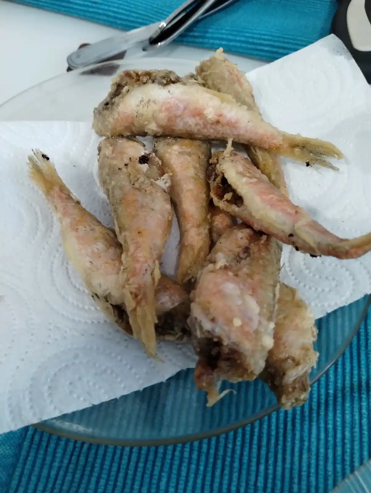 A close-up photo shows a pile of small, battered, and fried fish on a white paper towel, which is on a clear glass plate…