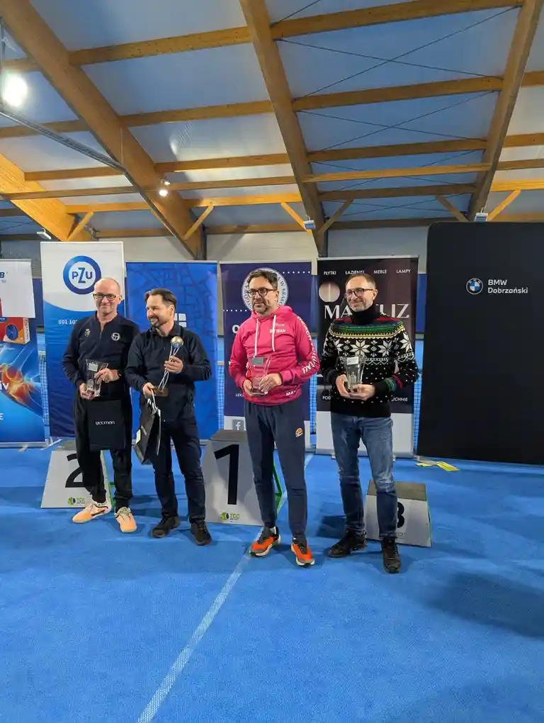 Four men stand on a podium with numbered steps (1, 2, 3) at an indoor sports event, each holding a trophy or award. The man…