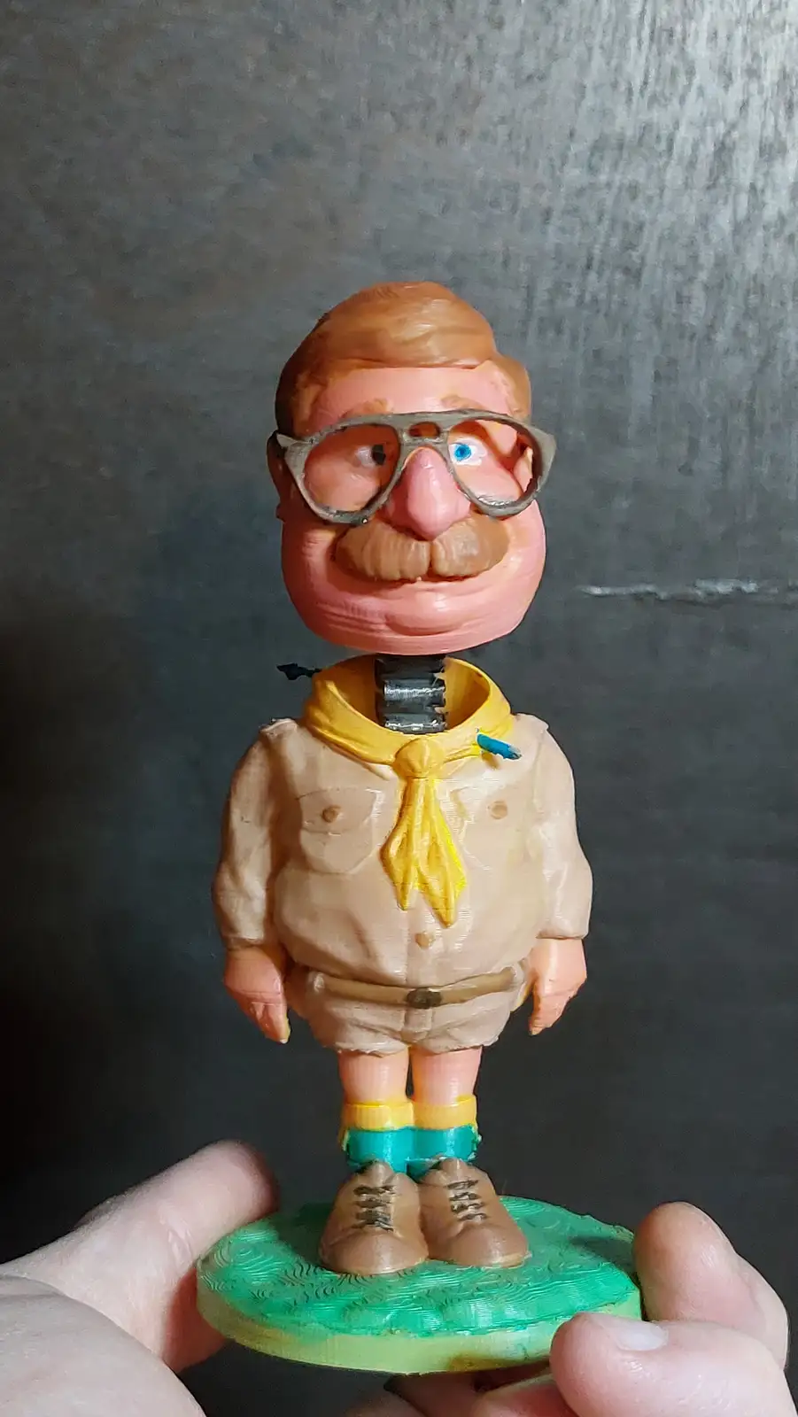 A person holds a 3D-printed bobblehead figurine of a man with glasses, a mustache, and a yellow neckerchief, wearing a beige…