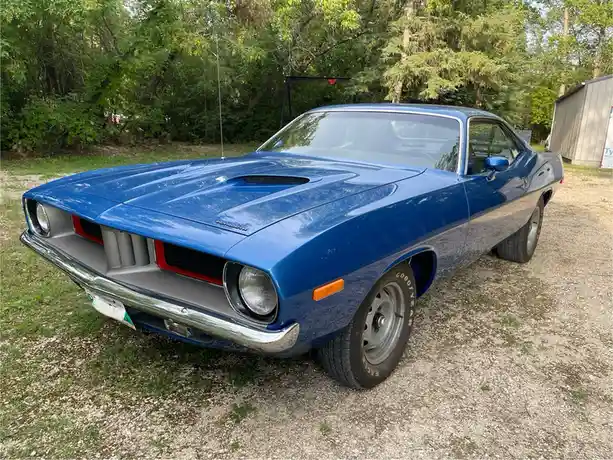 A blue 1970s Dodge Challenger with a hood scoop is parked on gravel in front of trees. The overlaid text reads 'i'm not a…