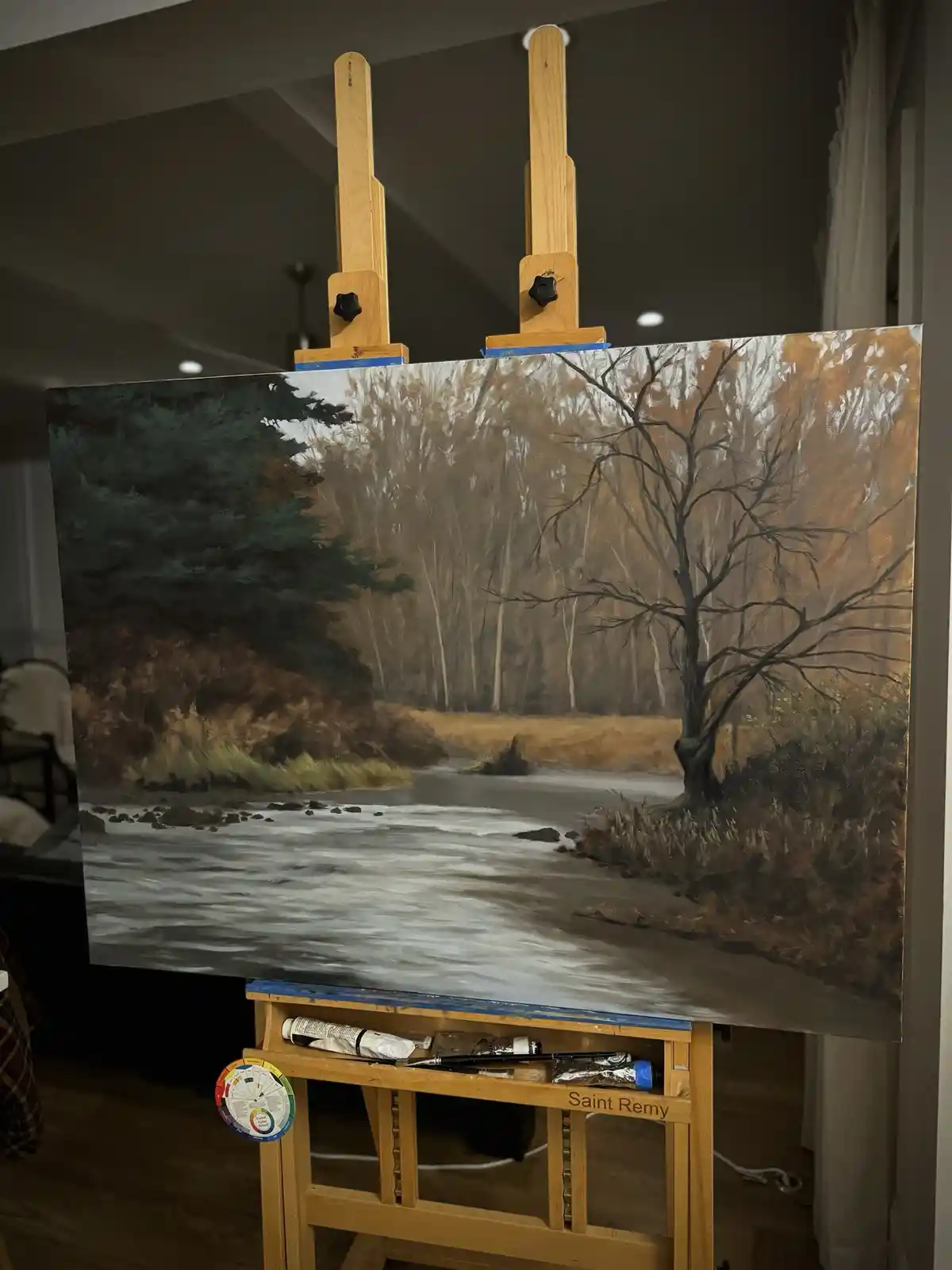 A large painting of a river with bare trees and autumnal foliage is displayed on a wooden easel in a room. The painting…