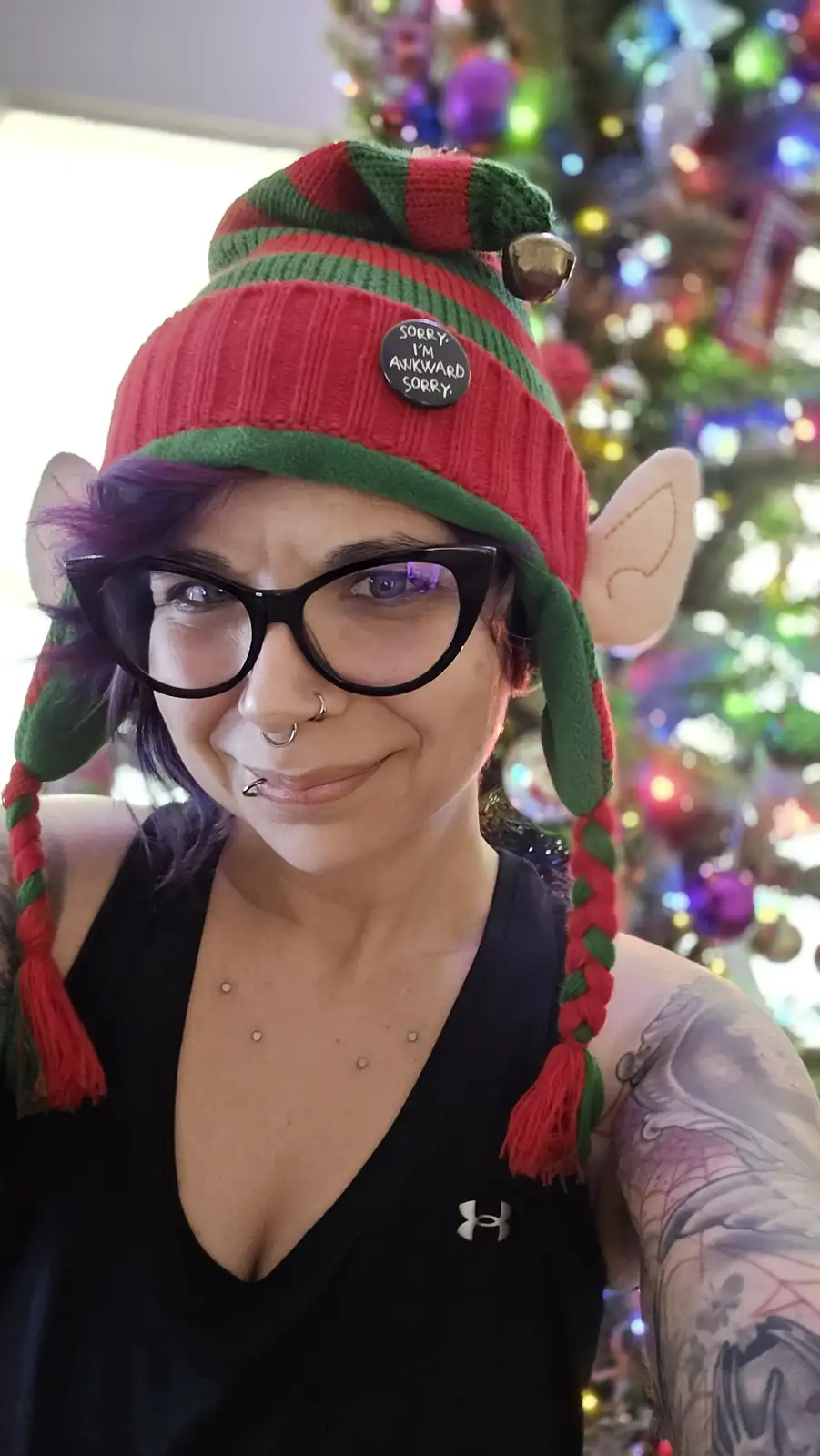 A person with purple hair, glasses, and multiple facial piercings wears a red-and-green striped elf hat with attached ears…