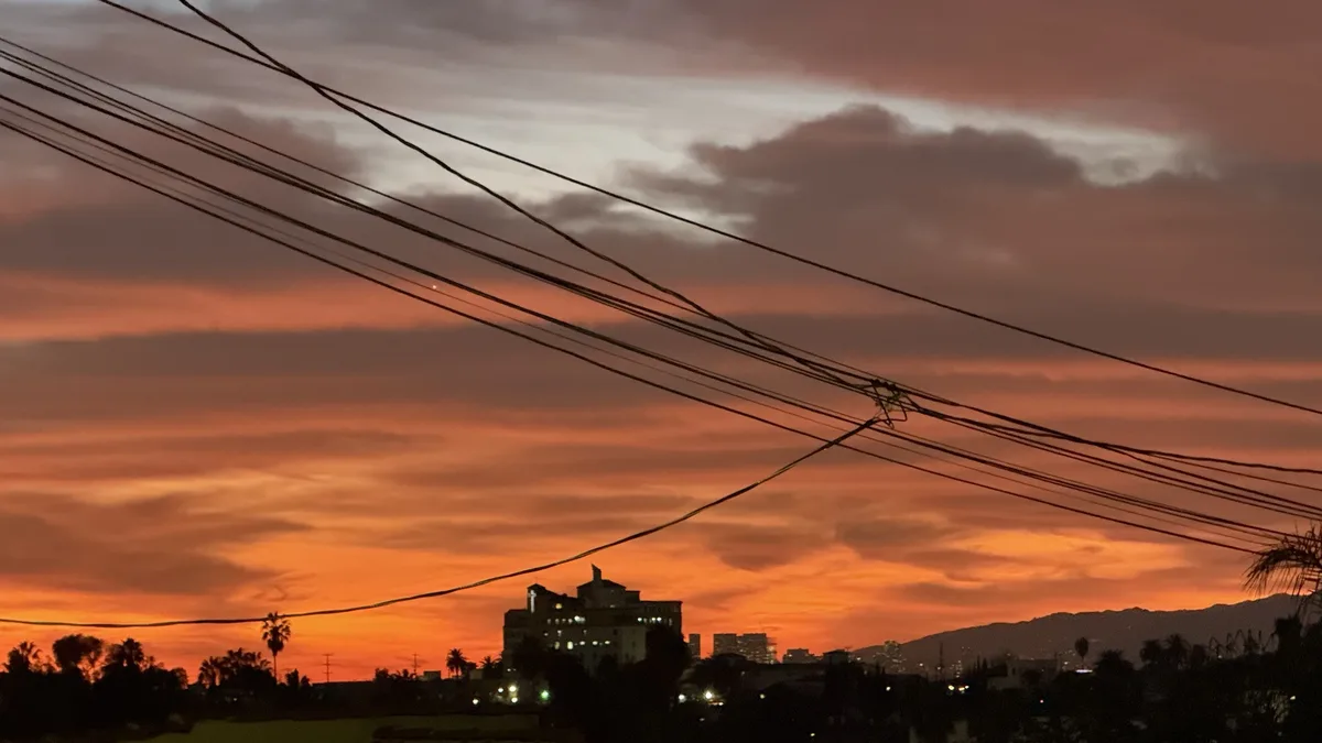 A dramatic sunset with orange and pink clouds, silhouetted buildings, and numerous power lines crossing the sky. The…
