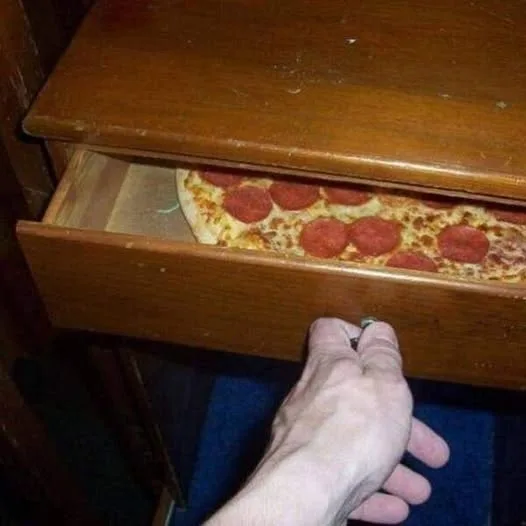 a person's hand is pulling open a wooden drawer to reveal a whole pepperoni pizza peeking out from inside. The drawer is…