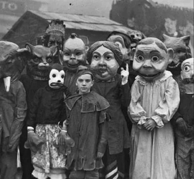 A black-and-white historical photo shows a group of children and adults wearing large, grotesque, handmade masks resembling…