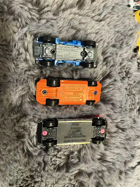 Three Hot Wheels toy cars are shown upside down on a gray furry surface, revealing their undersides with visible…
