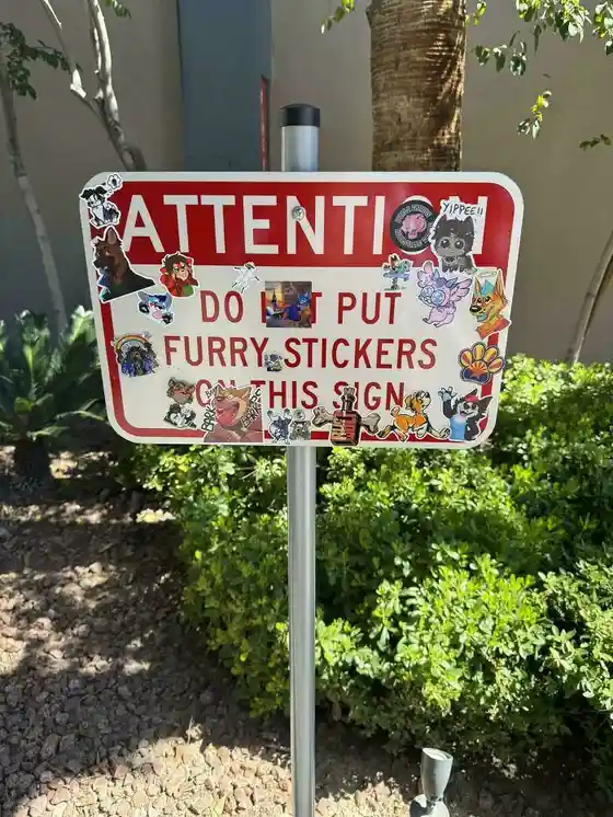A red and white warning sign, partially obscured by numerous colorful furry-themed stickers, reads 'ATTENTI DO NOT PUT FURRY…