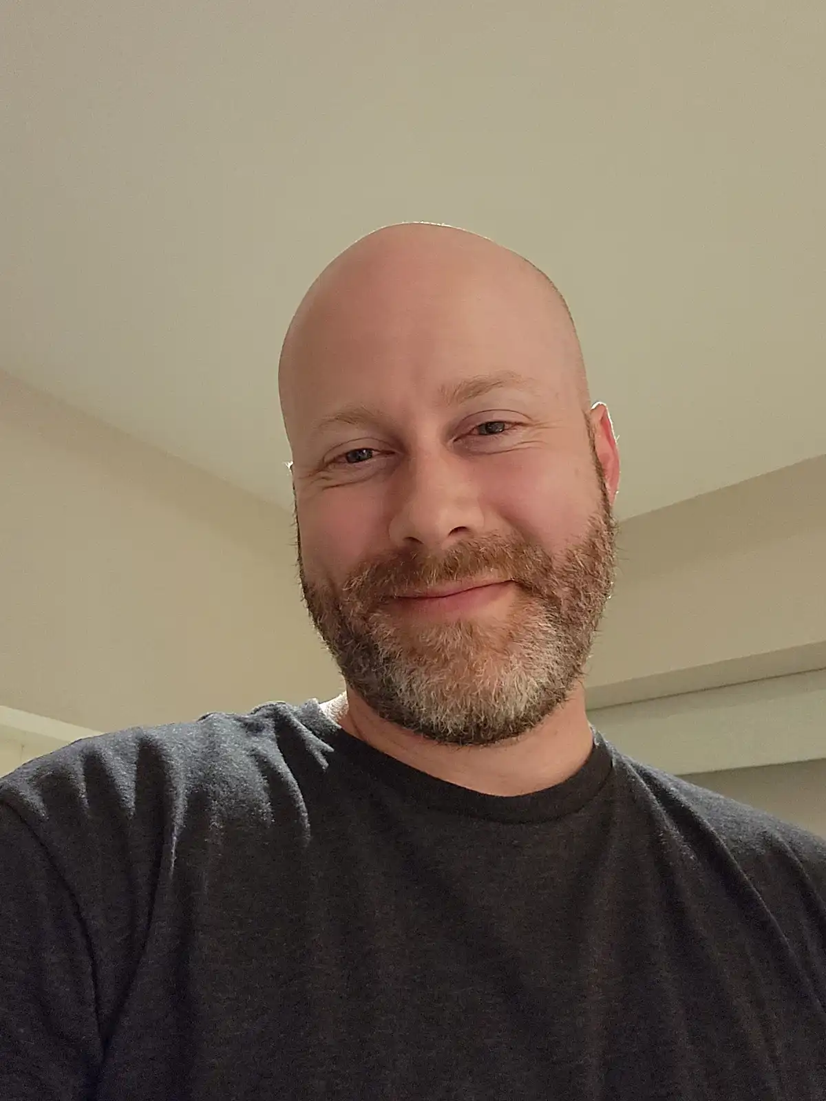 A bald man with a full beard and mustache, wearing a dark gray t-shirt, smiles at the camera. The overlaid text reads 'i…