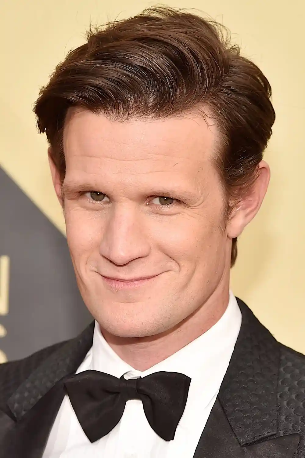 A close-up photo of actor Matt Smith in a black tuxedo with a bow tie, smiling subtly at the camera. The overlaid text reads…