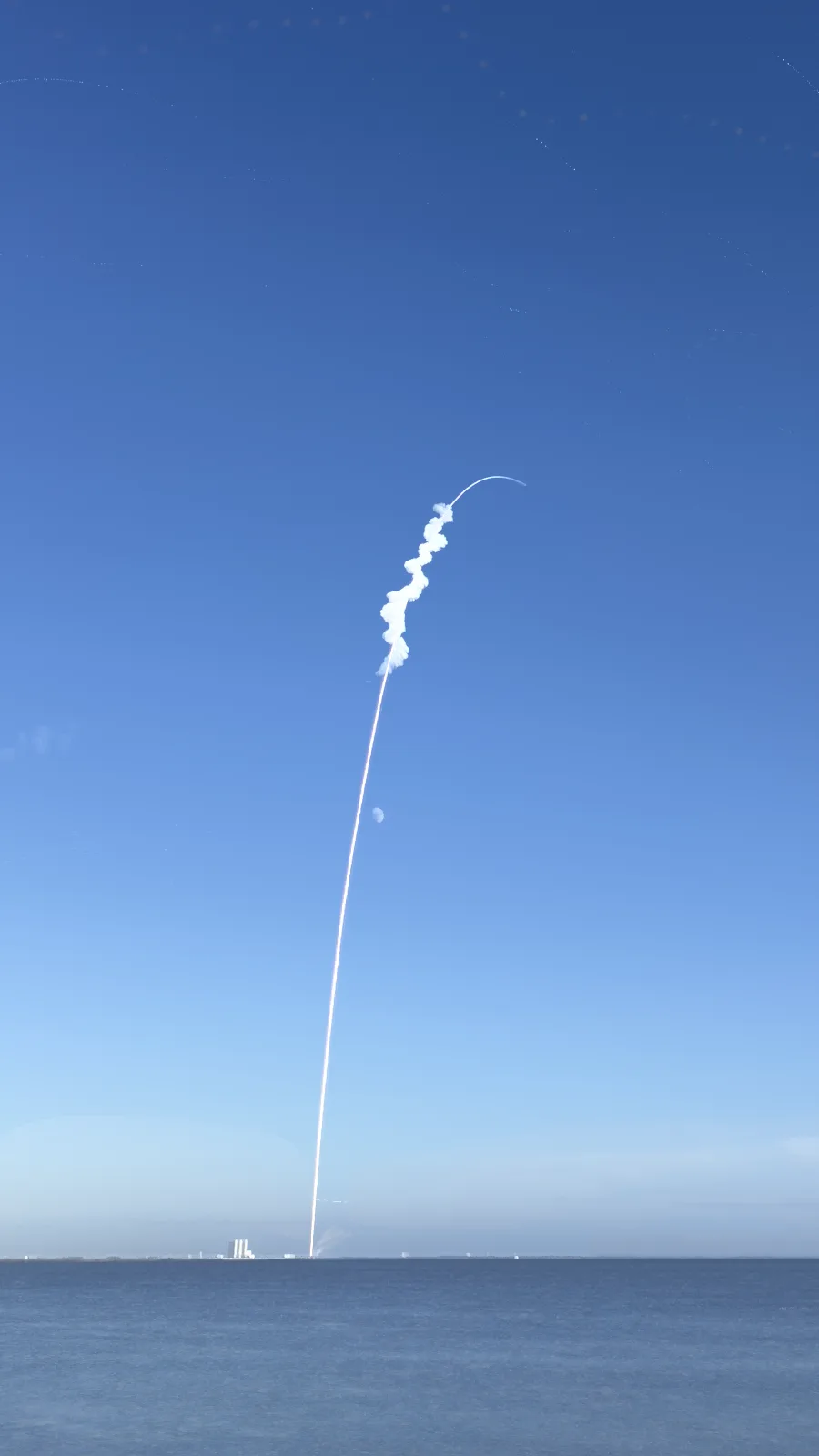 A rocket launches vertically over a body of water under a clear blue sky, leaving a bright white exhaust trail that curves…