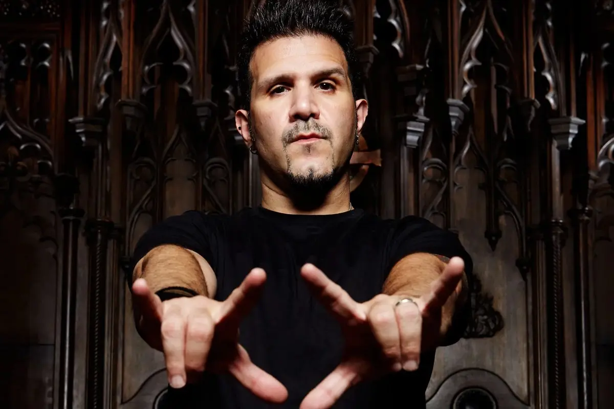 A man with a goatee and spiky hair, wearing a black t-shirt, stands in front of ornate dark wood paneling. He points both…
