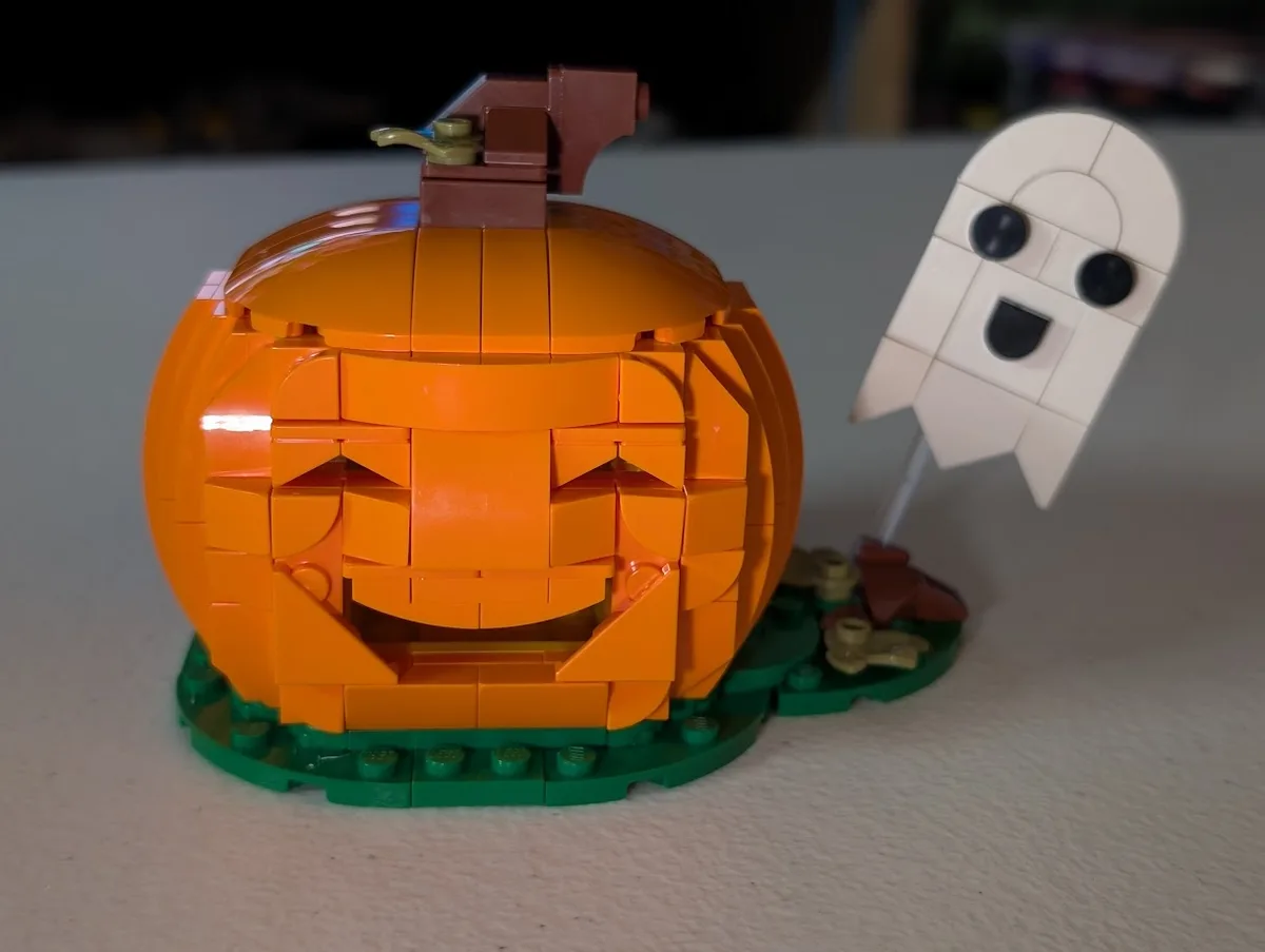 A close-up photo of a LEGO pumpkin with a carved jack-o'-lantern face, sitting on a green baseplate, next to a small white…