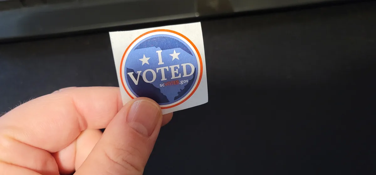 A hand holds a circular 'I VOTED' sticker featuring a blue background with a white silhouette of South Carolina, two white…