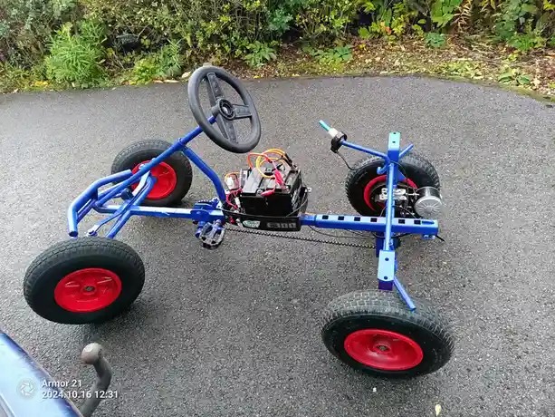 A blue, four-wheeled go-kart with red wheels and a black steering wheel sits on an asphalt path, visibly missing a seat. The…