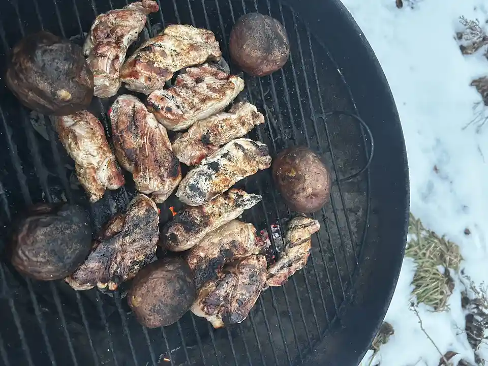 An overhead shot shows a black charcoal grill with numerous pieces of meat and several whole potatoes cooking over glowing…