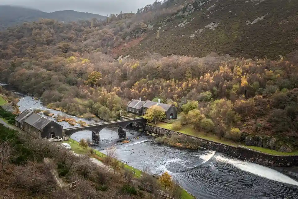 An aerial view of a scenic river valley with stone buildings, a bridge, and autumn-colored trees on hillsides. The image has…