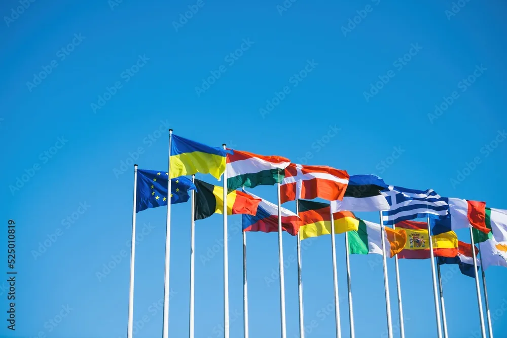 A row of international flags, including Ukraine and the European Union, waving against a clear blue sky. The overlaid text…